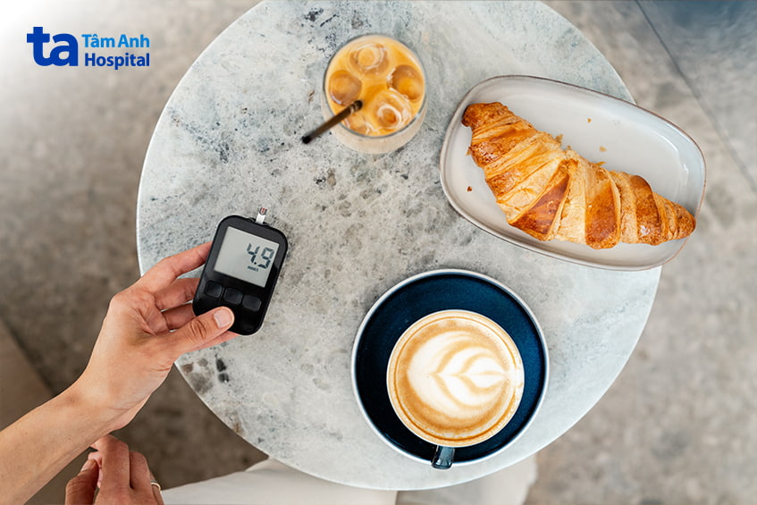 Người cầm máy đo đường huyết bên ly cà phê và bánh croissant trên bàn đá