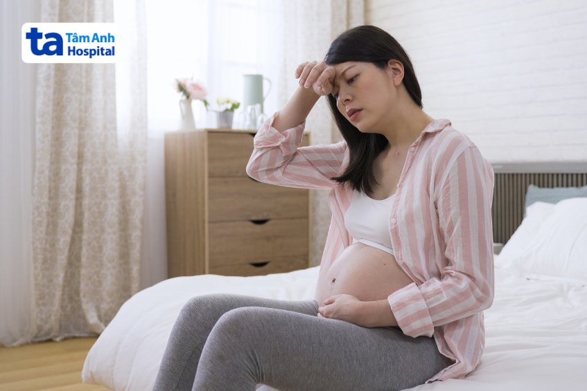 Bà bầu mệt mỏi ngồi trên giường, tay ôm bụng và áp trán thể hiện dấu hiệu khó chịu thai kỳ