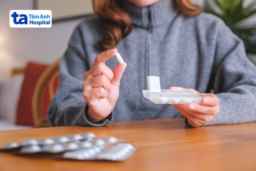 Người phụ nữ cầm viên thuốc màu trắng với hộp chia liều trên bàn