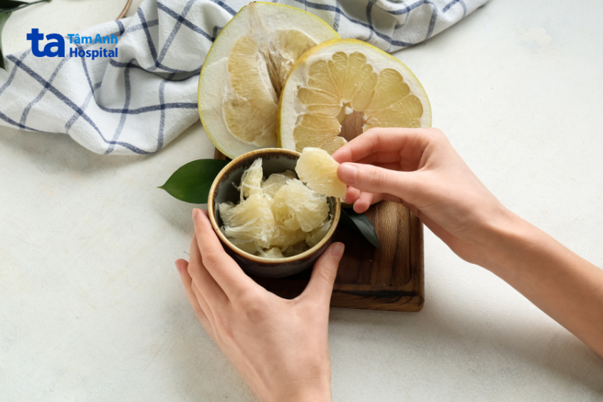 Tay người đang lấy múi bưởi trong chén, phía sau là quả bưởi bổ đôi dùng minh họa mẹ bầu ăn bưởi