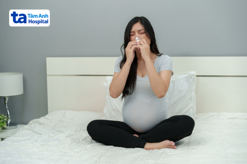 Bà bầu bị cảm cúm, ngồi trên giường và dùng khăn giấy lau mũi