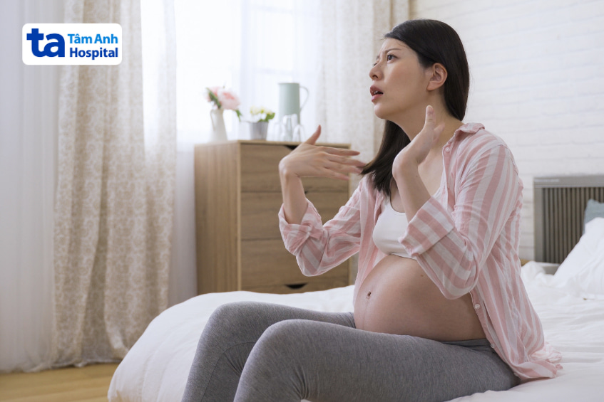 Bà bầu ngồi trên giường, biểu cảm khó chịu do nóng bức khi mang thai.