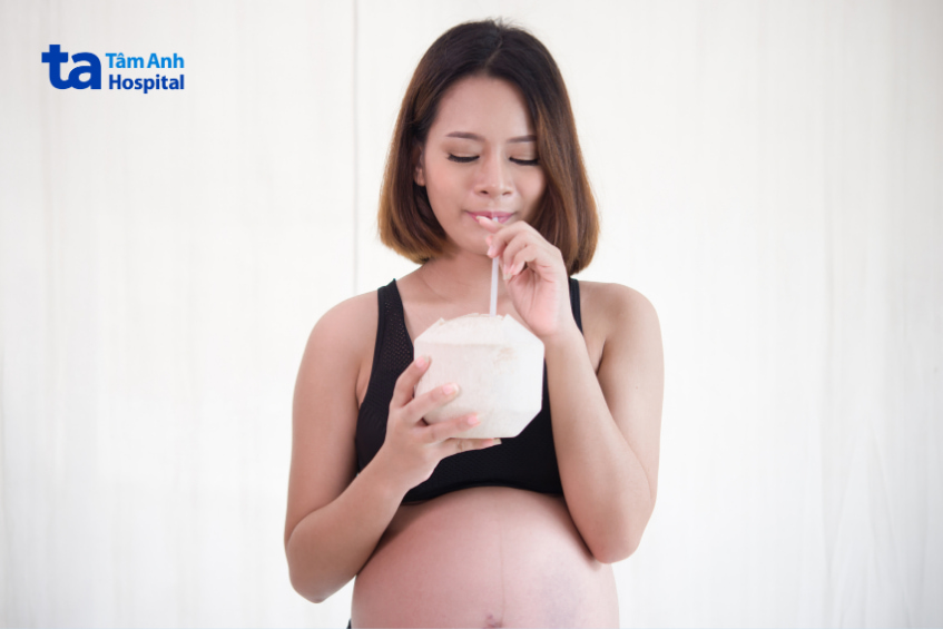 Bà bầu mặc áo thể thao đen đang uống nước dừa tươi bằng ống hút trong phòng sáng