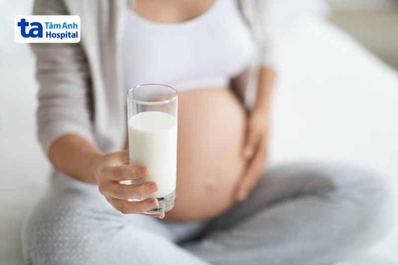Bà bầu cầm ly sữa ngồi trên giường, chăm sóc sức khỏe thai kỳ bằng dinh dưỡng lành mạnh