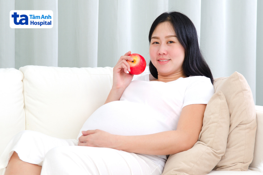 Phụ nữ mang thai mặc đồ trắng đang ăn táo đỏ trên ghế sofa