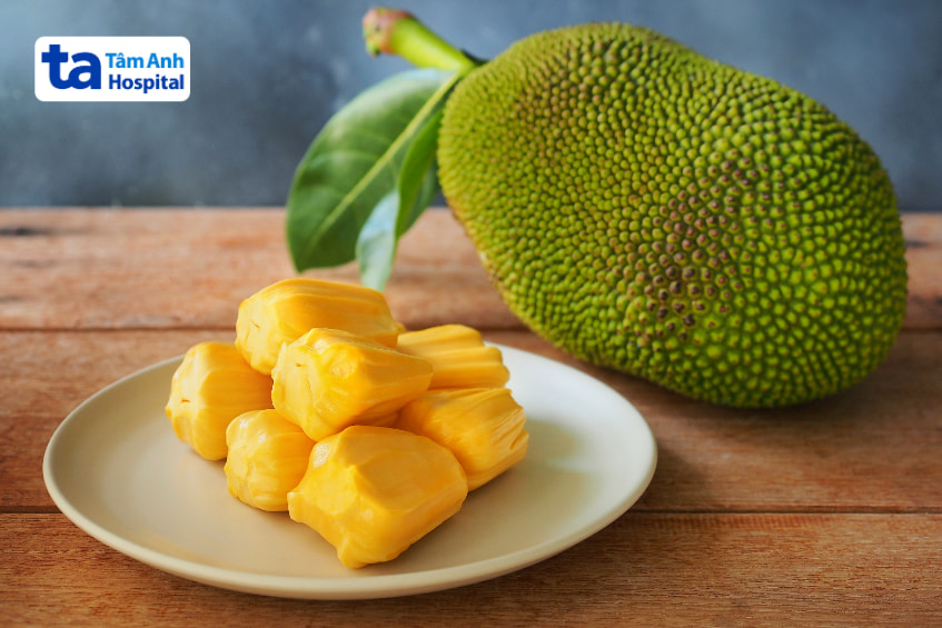 Dĩa mít chín đặt trước trái mít xanh nguyên vỏ trên nền gỗ.