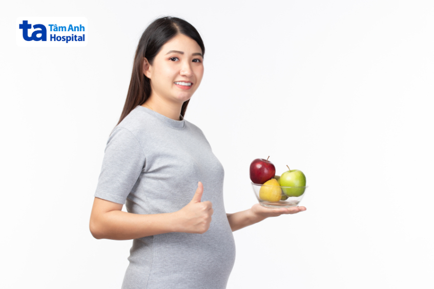 Mẹ bầu cười tươi giơ ngón tay cái, tay cầm tô trái cây gồm táo và lê