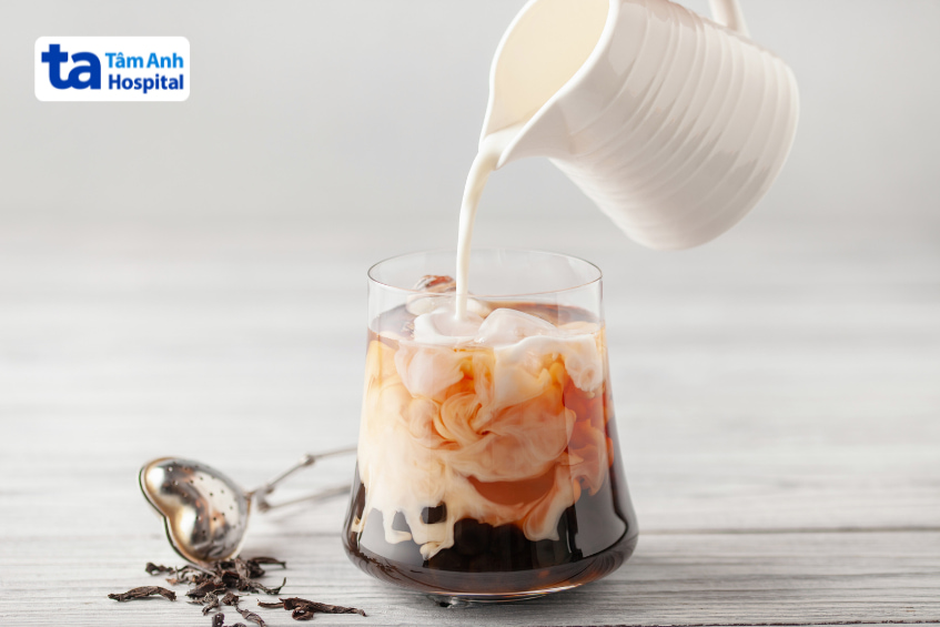 Cận cảnh rót sữa vào trà đen có đá, tạo thành trà sữa hòa quyện, bên cạnh là lá trà khô và thìa lọc